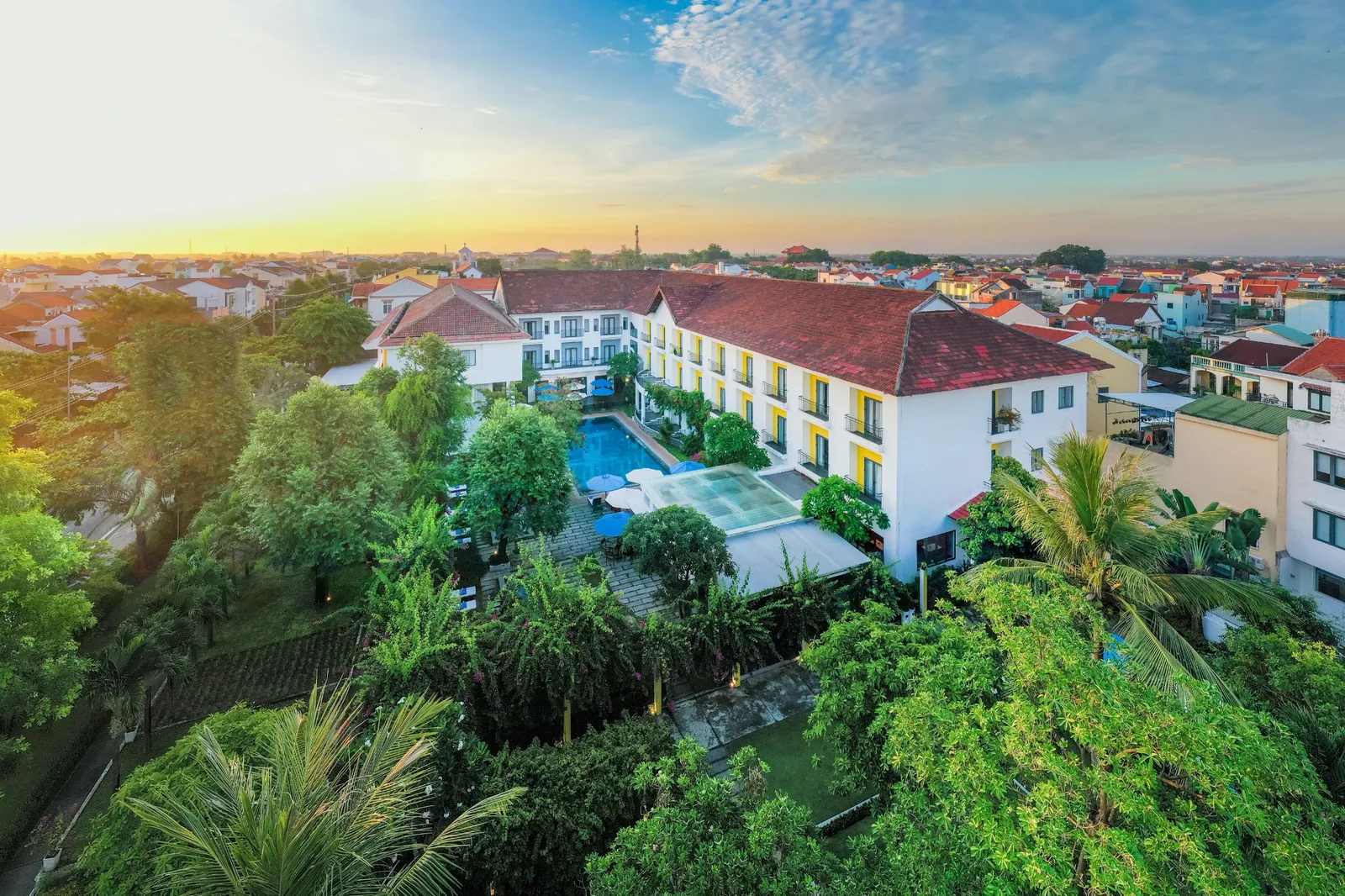 Ảnh flycam EMM Hotel Hoi An lúc bình minh: toà nhà trắng mái ngói đỏ điểm vàng, hồ bơi xanh giữa vườn cây, xung quanh là mái nhà Hội An