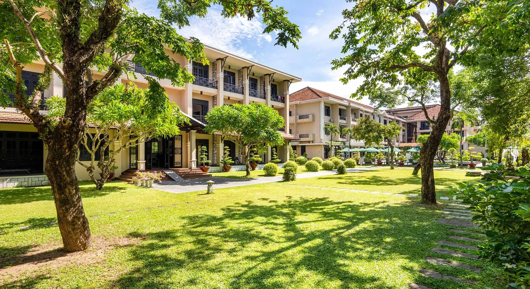 Bãi cỏ rộng và hàng cây cổ thụ trước các dãy nhà ba tầng màu kem kiến trúc Đông Dương của Hoi An Historic Hotel