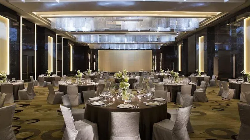 Sảnh tiệc JW Marriott Hotel Hanoi bày bàn tròn phủ khăn nâu, ghế bọc vải xám, đèn chùm pha lê dài trên trần