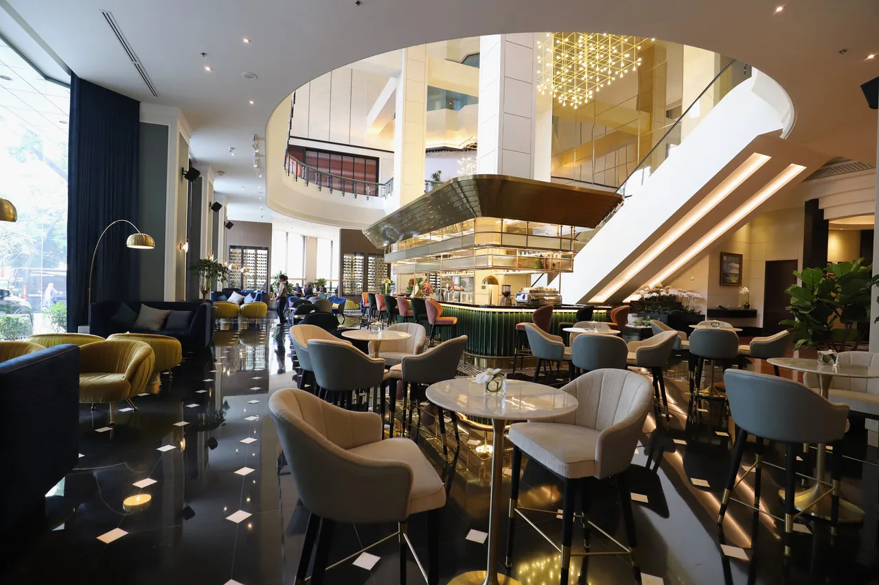 Sảnh lounge Meliá Hanoi với ghế bọc nỉ xám, sàn đá đen bóng, quầy bar và cầu thang lượn lên tầng trên
