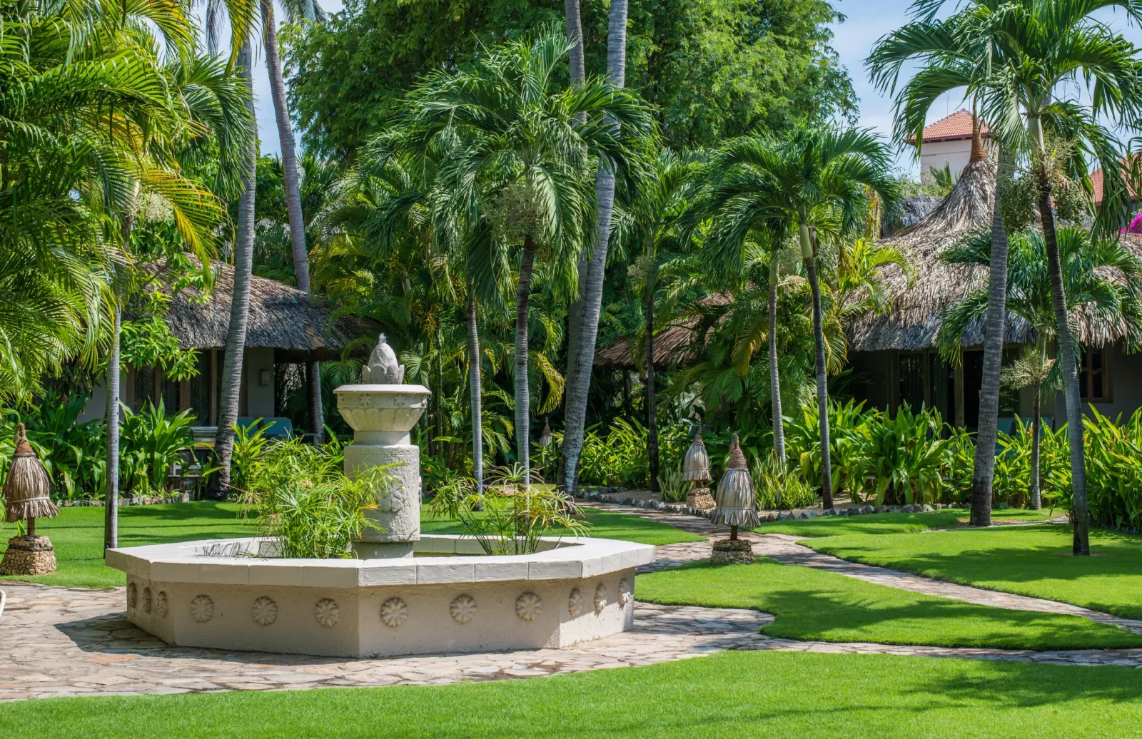 Đài phun nước giữa bãi cỏ và các bungalow mái lá khuất dưới hàng dừa tại Mia Resort Mui Ne