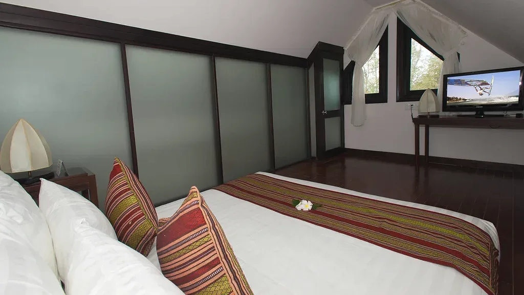 Phòng ngủ trên gác lửng trần dốc tại Muine Bay Resort, giường lớn phủ khăn thổ cẩm đỏ, cửa sổ nhỏ và tivi đặt sát tường