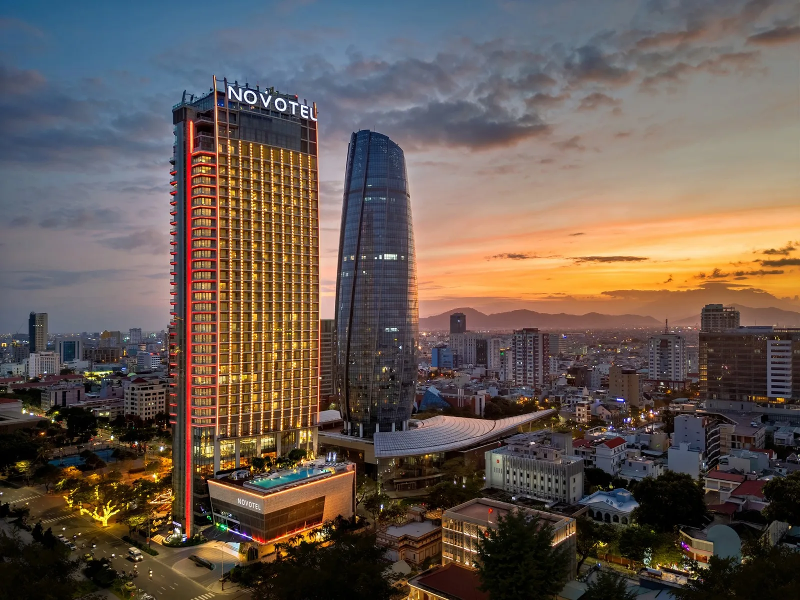 Tòa tháp Novotel Danang Premier Han River lên đèn lúc hoàng hôn giữa trung tâm Đà Nẵng, phía xa là dãy núi