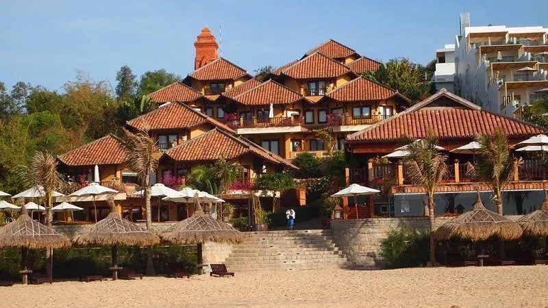 Poshanu Resort với cụm nhà mái ngói đỏ kiến trúc Chăm và tháp gạch phía sau, bậc thang dẫn xuống bãi cát có dù lá