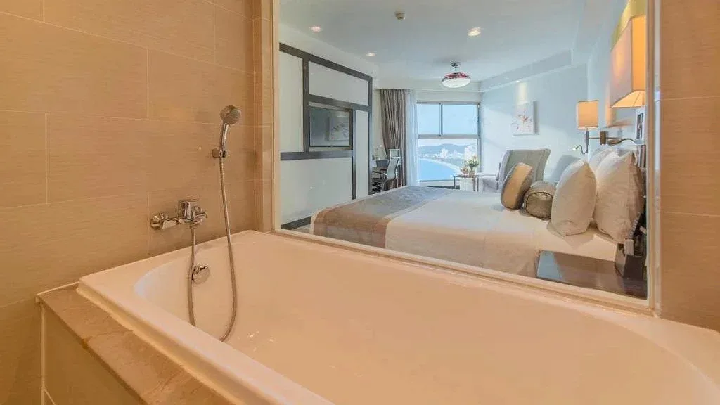 Bồn tắm trắng sát vách kính trong phòng Premier Havana Nha Trang, nhìn qua giường ra cửa sổ thấy bờ biển
