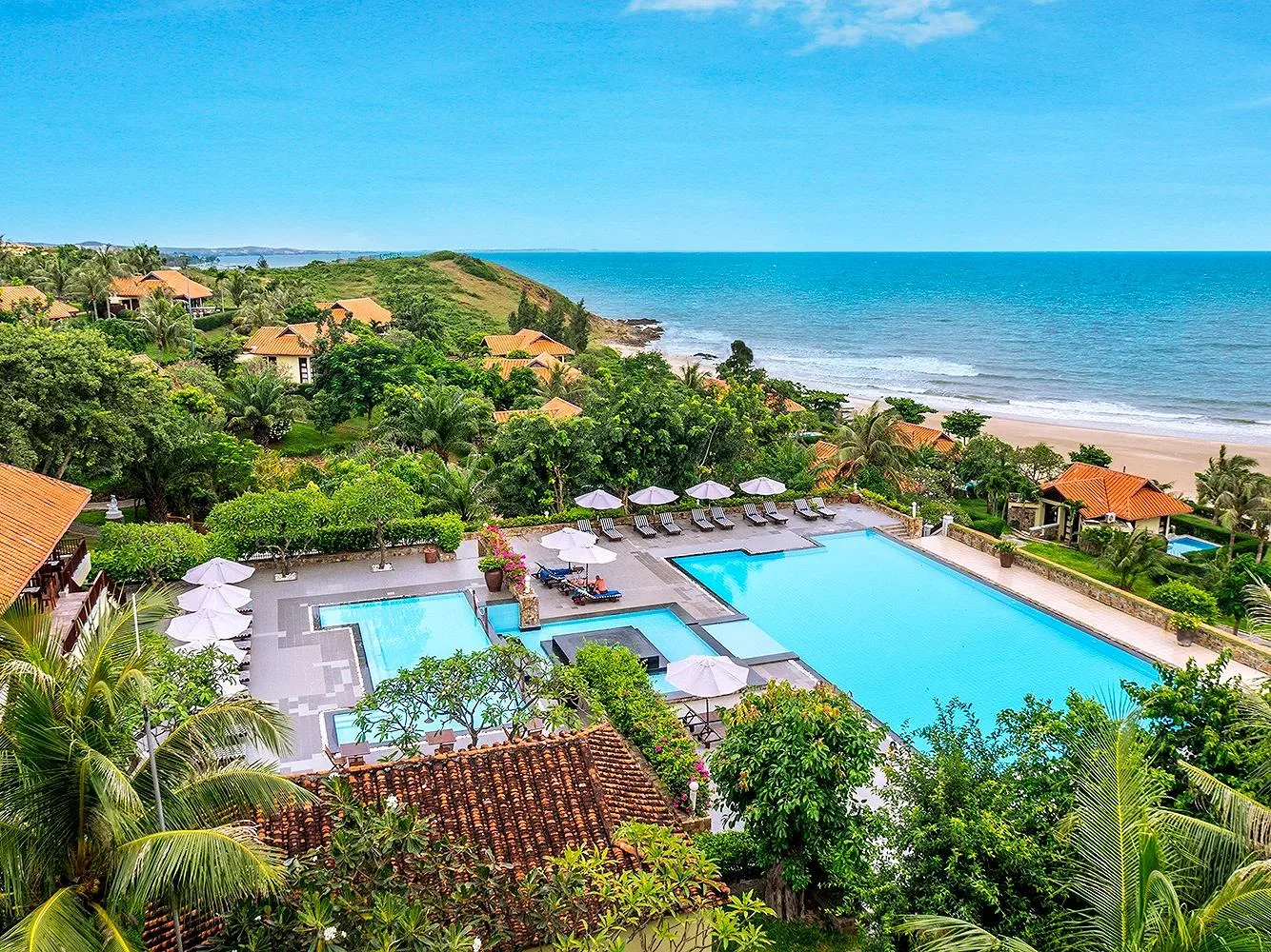 Romana Resort & Spa Phan Thiết nhìn từ trên đồi: cụm hồ bơi xanh, villa mái ngói đỏ giữa vườn cây và bãi cát vàng chạy dọc biển