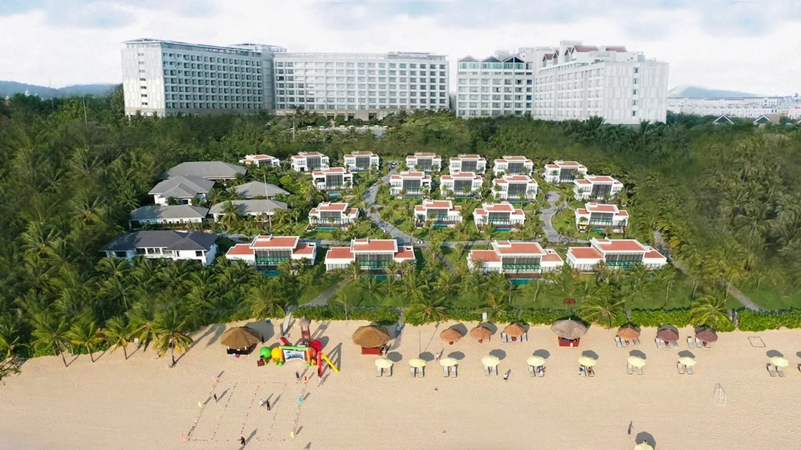 Toàn cảnh khu villa mái đỏ và khối khách sạn phía sau, hàng dù và khu vui chơi trẻ em trên bãi cát